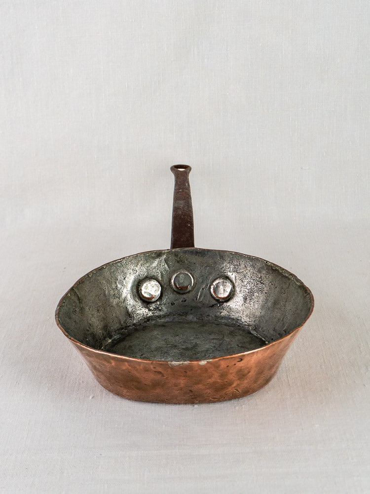 Antique French copper sauté pan with upright handle and three interior circular indents, plain backdrop.