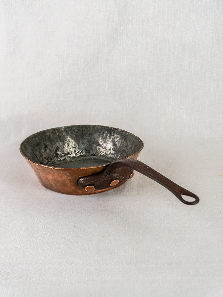 Antique 19th-century French copper sauté pan with dark handle and tarnished interior on light fabric.