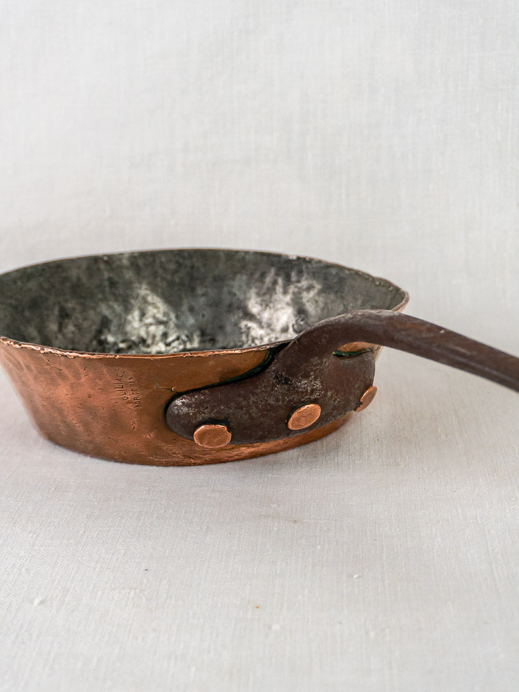 Antique 19th-century French copper sauté pan with dark metal handle and tarnished interior on fabric.