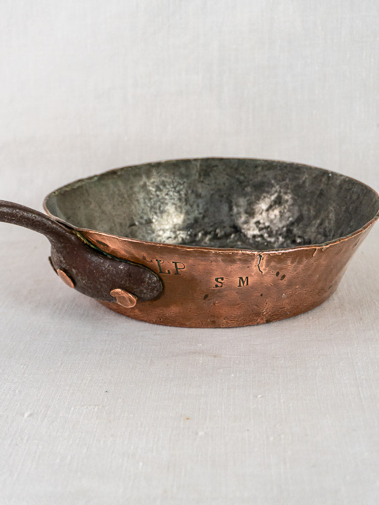 Antique 19th-century French copper sauté pan with dark metal handle and engraved initials, plain background.
