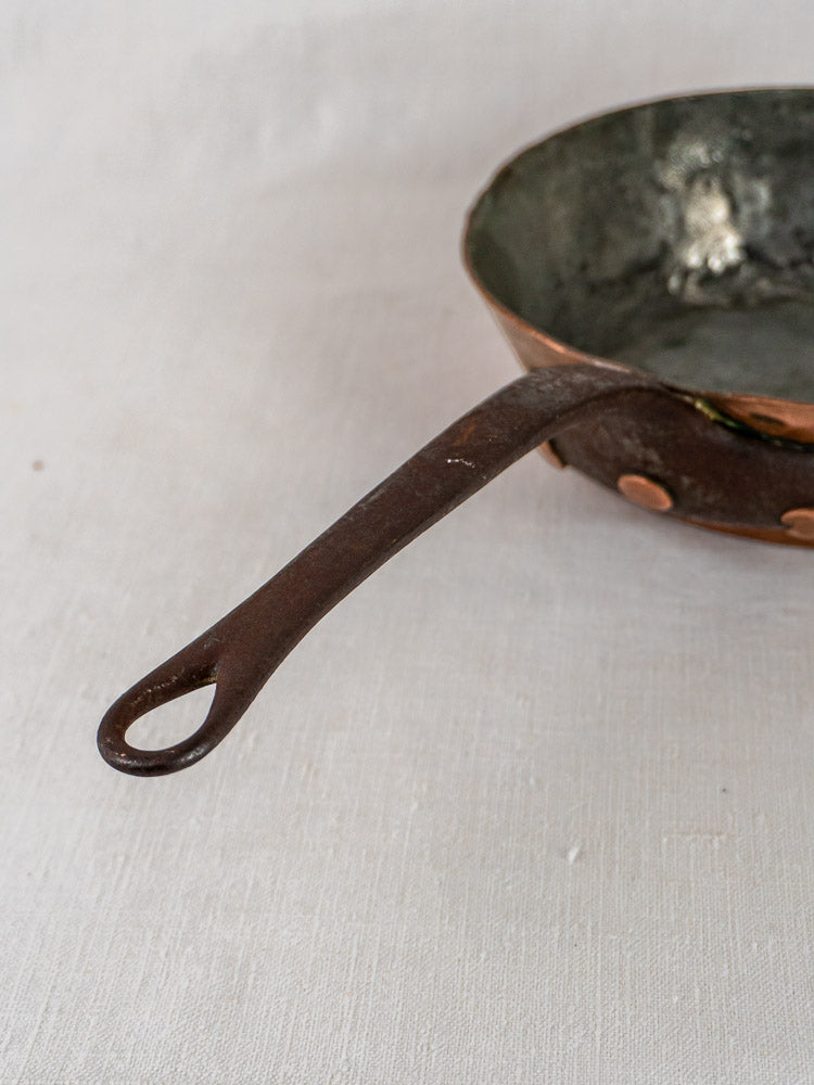 Antique 19th-century French copper sauté pan with riveted long handle on light surface, hand-hammered craftsmanship.