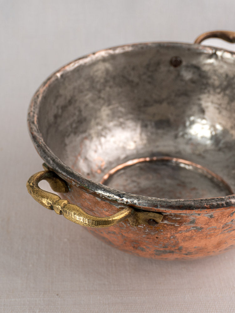 Antique French copper bowl with brass handles and worn silvery interior on light fabric surface.