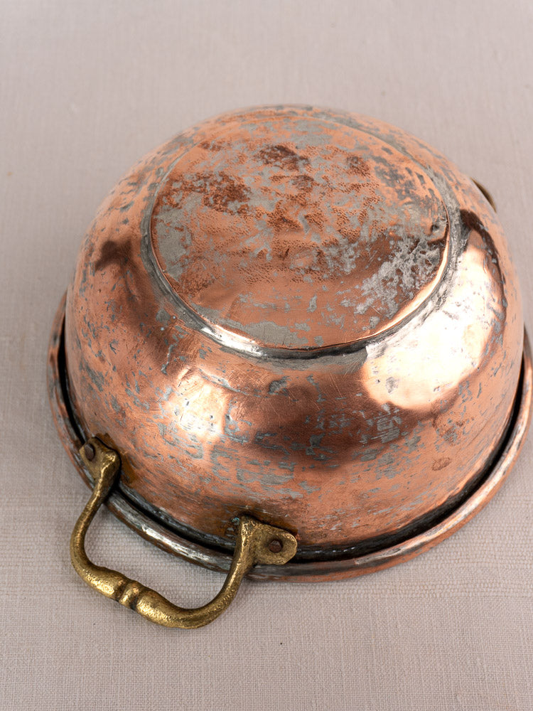 Antique French copper bowl with brass handles, displaying rich patina, shown upside-down on a light textured surface.