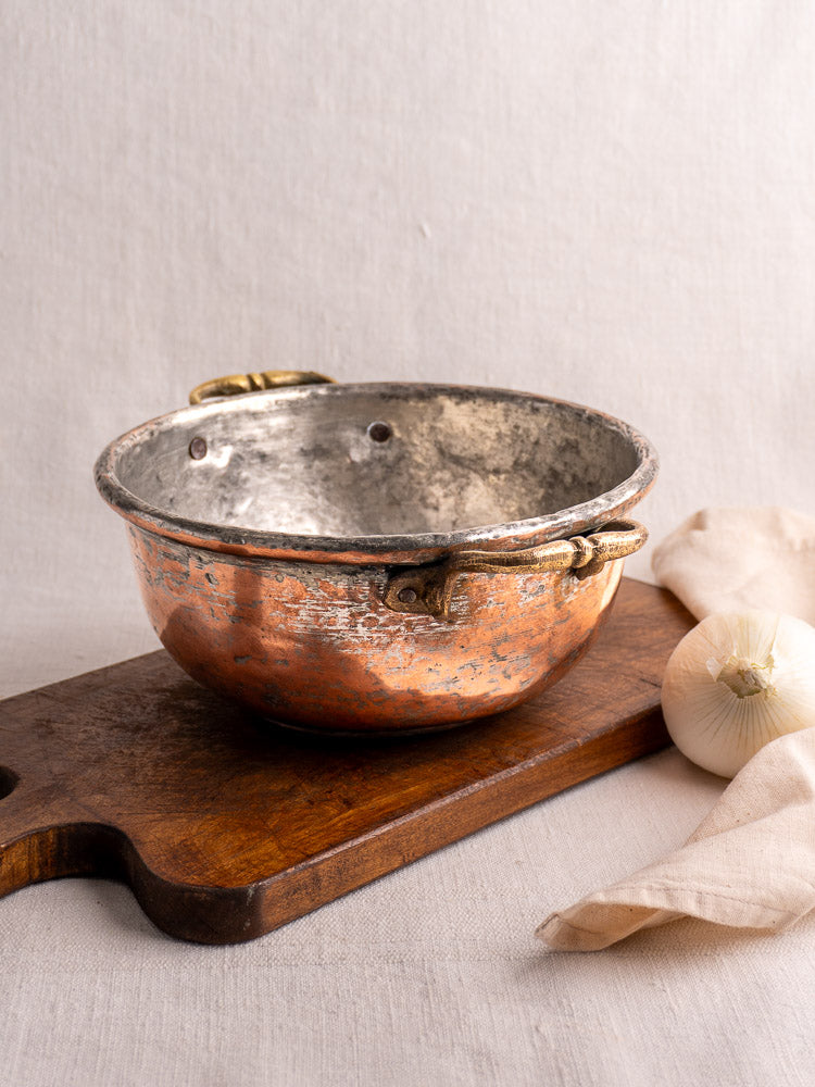 Antique French copper bowl with brass handles on a wooden board beside onion and beige cloth, softly lit neutral background.