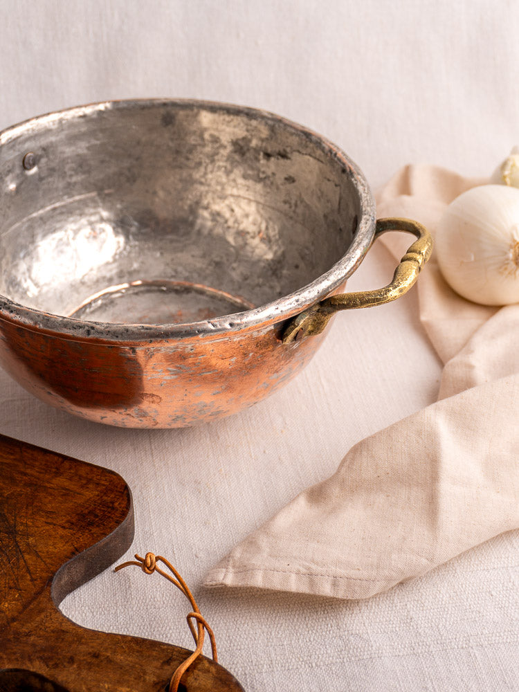 Antique French copper bowl with brass handles on cream cloth, beside white onions and a wooden cutting board.