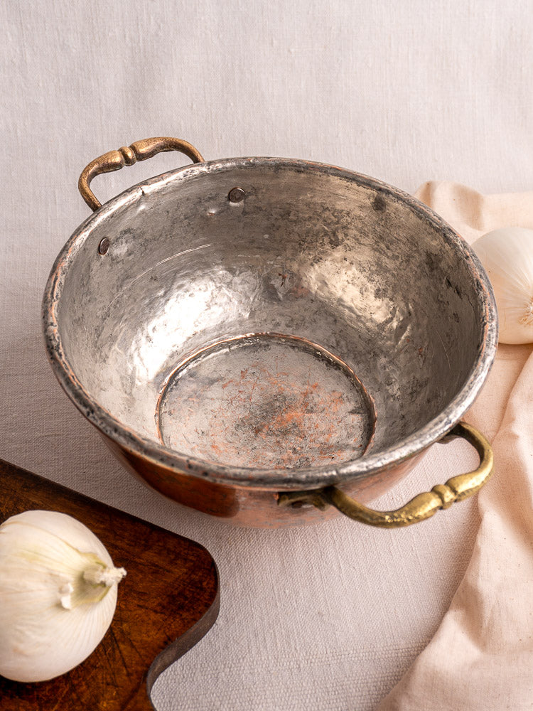 Antique French copper bowl with brass handles on light fabric, flanked by two garlic bulbs and a wooden board.