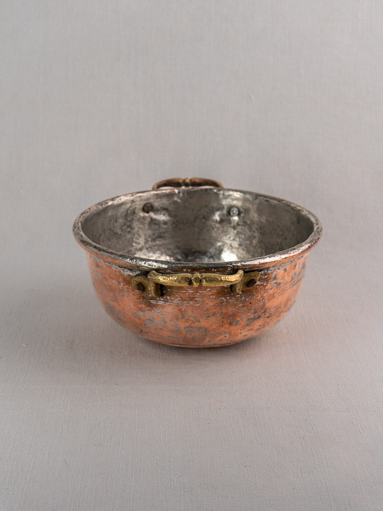 Antique French copper bowl with silvery interior and brass handles on a light gray fabric surface.