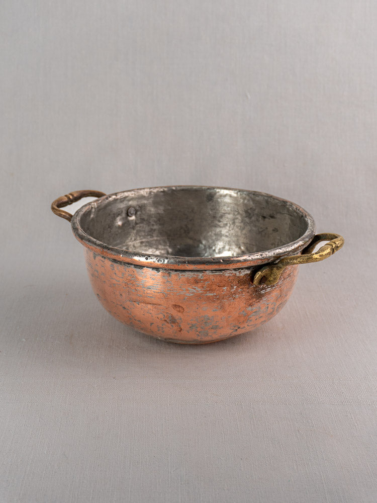 Antique French copper bowl with brass handles and worn silvery interior on a light gray background.