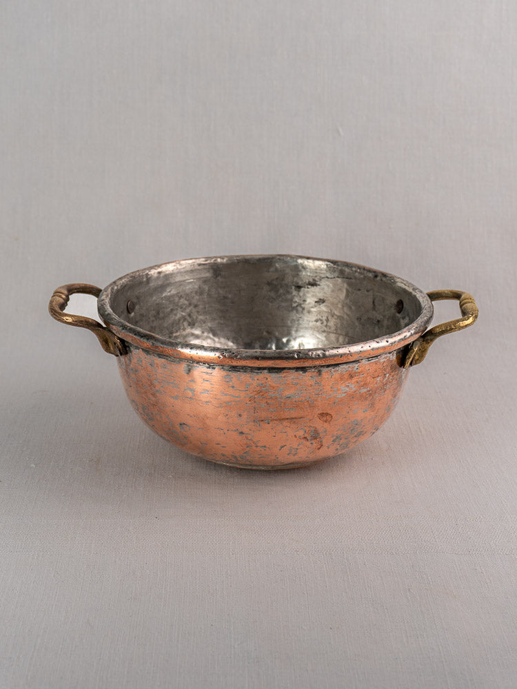 Antique French hammered copper bowl with brass handles, worn silver interior, on neutral fabric background.