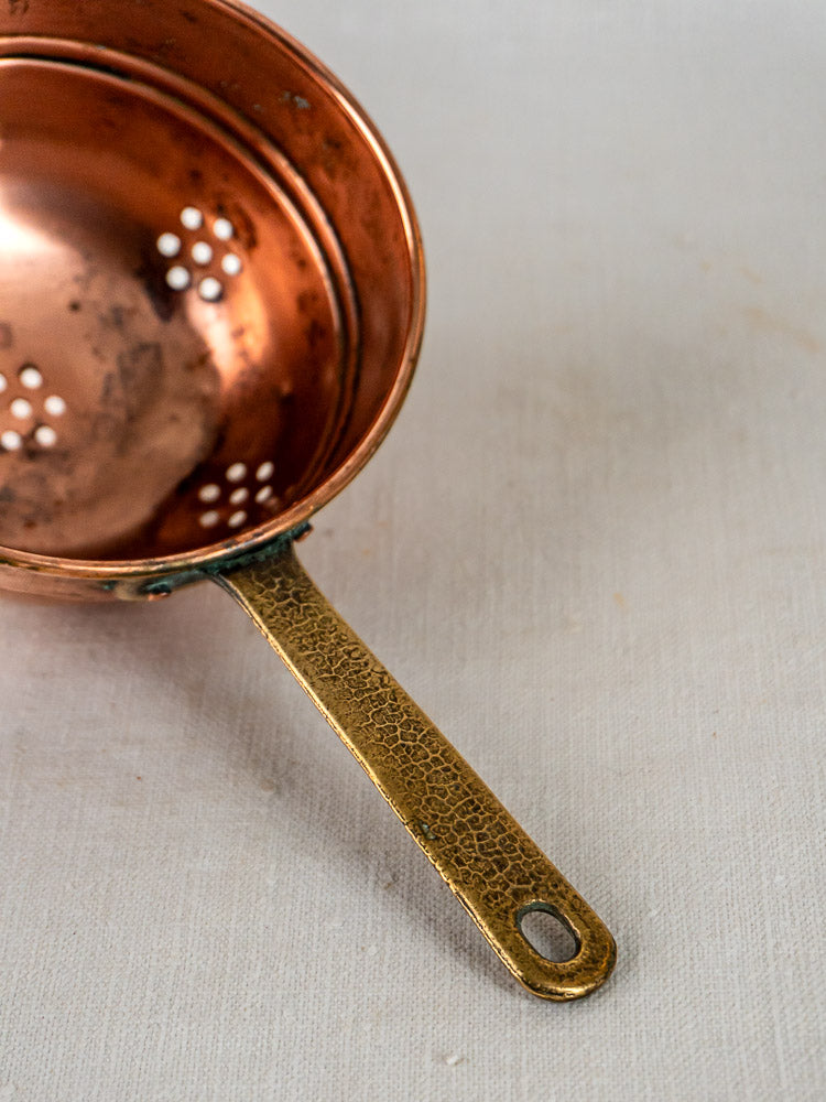 Antique French copper strainers trio with textured brass handles on white fabric background.
