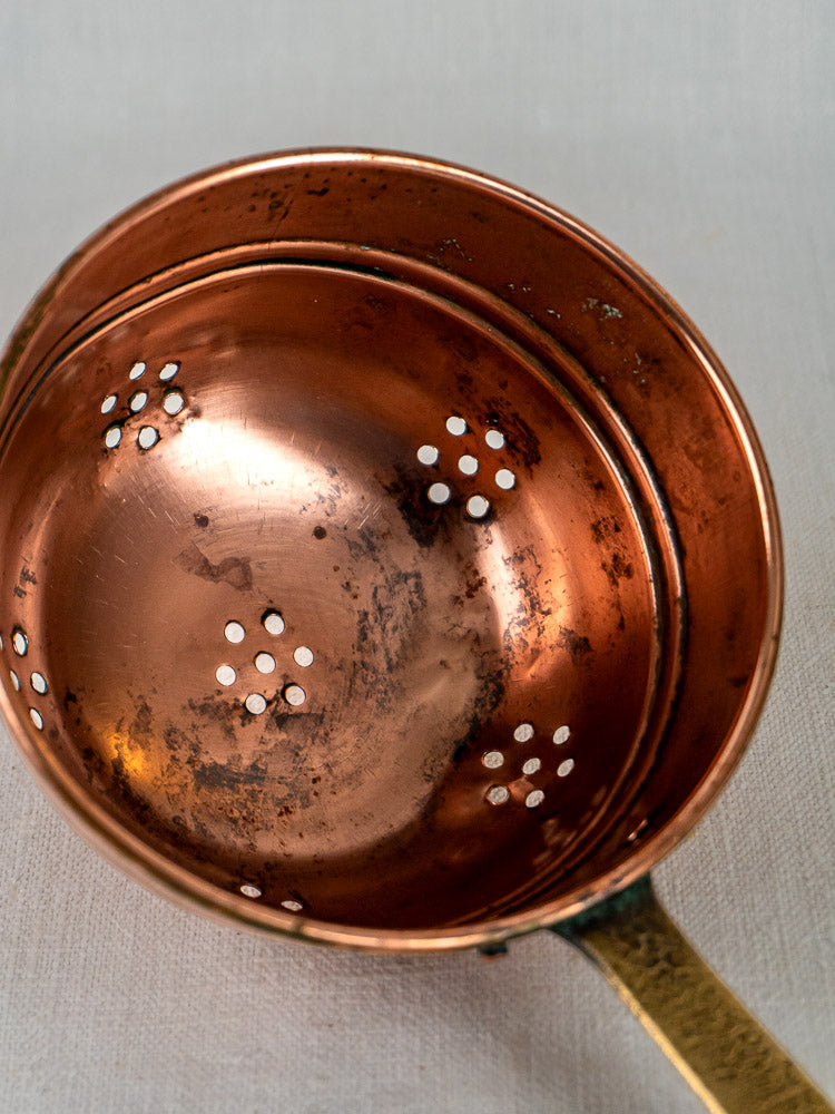Antique French copper strainer with brass handle, classic perforations, on light fabric background.