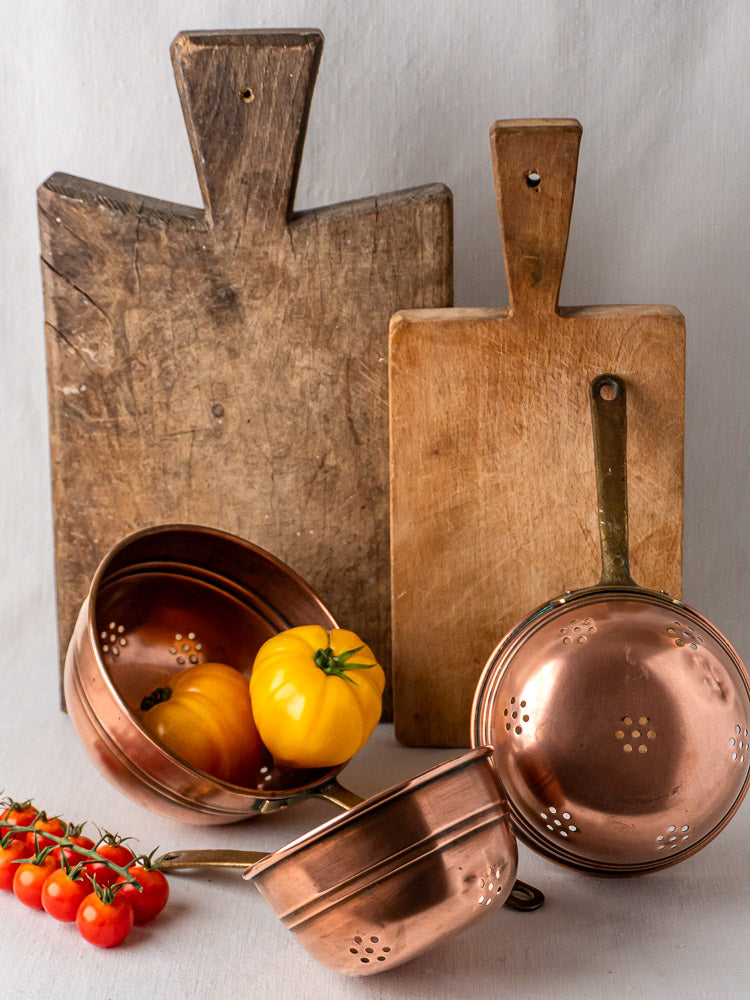 Antique French copper strainers with brass handles, one holding yellow tomatoes; wooden boards behind.