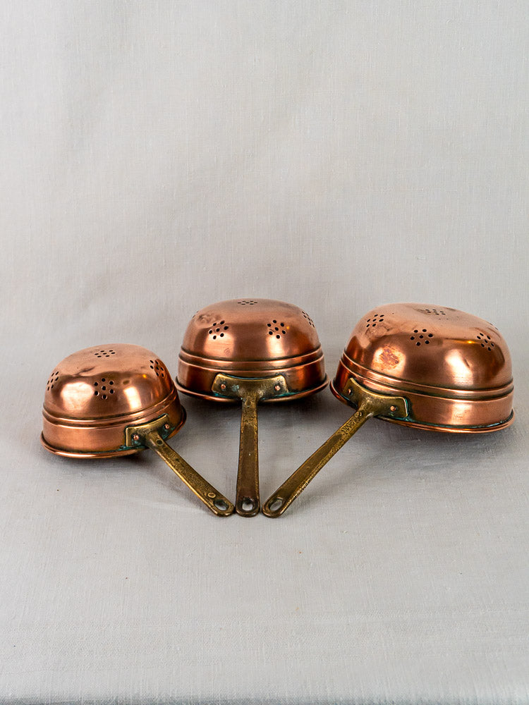 Antique French copper strainer set with brass handles, arranged upside down on neutral fabric.