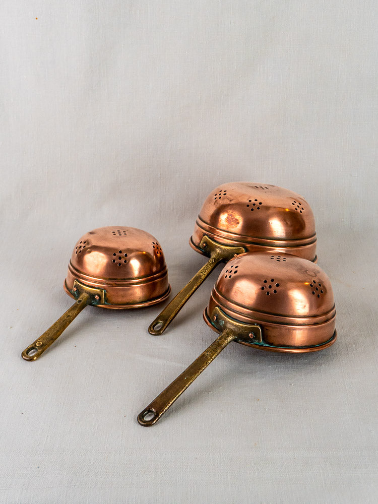 Antique French copper strainers with brass handles, graduated set of three on light gray fabric.