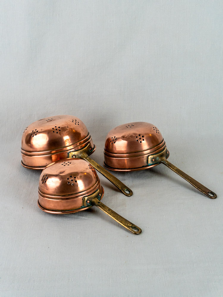 Antique French copper strainers, graduated trio with brass handles and perforated lids, on gray surface.