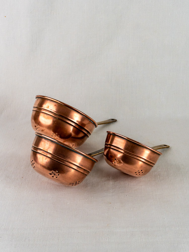 Antique French copper strainers with brass handles, graduated set of three on white fabric background.