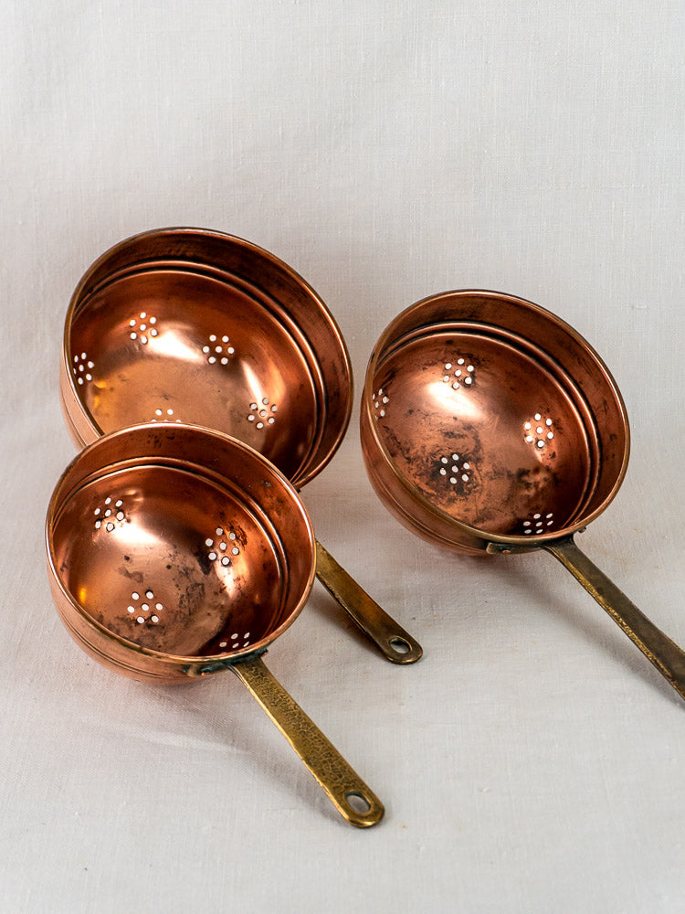 Antique French copper strainer set with brass handles, arranged on a light fabric background.