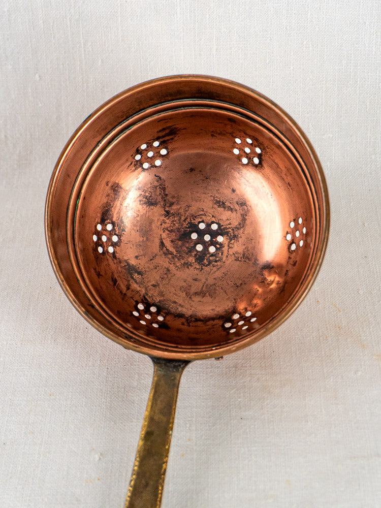 Antique French copper strainer trio with brass handles, decorative holes, on light fabric background.