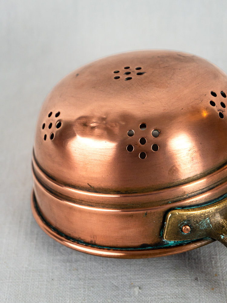 French copper strainer with decorative perforations and brass handle from antique graduated set, on light gray background.