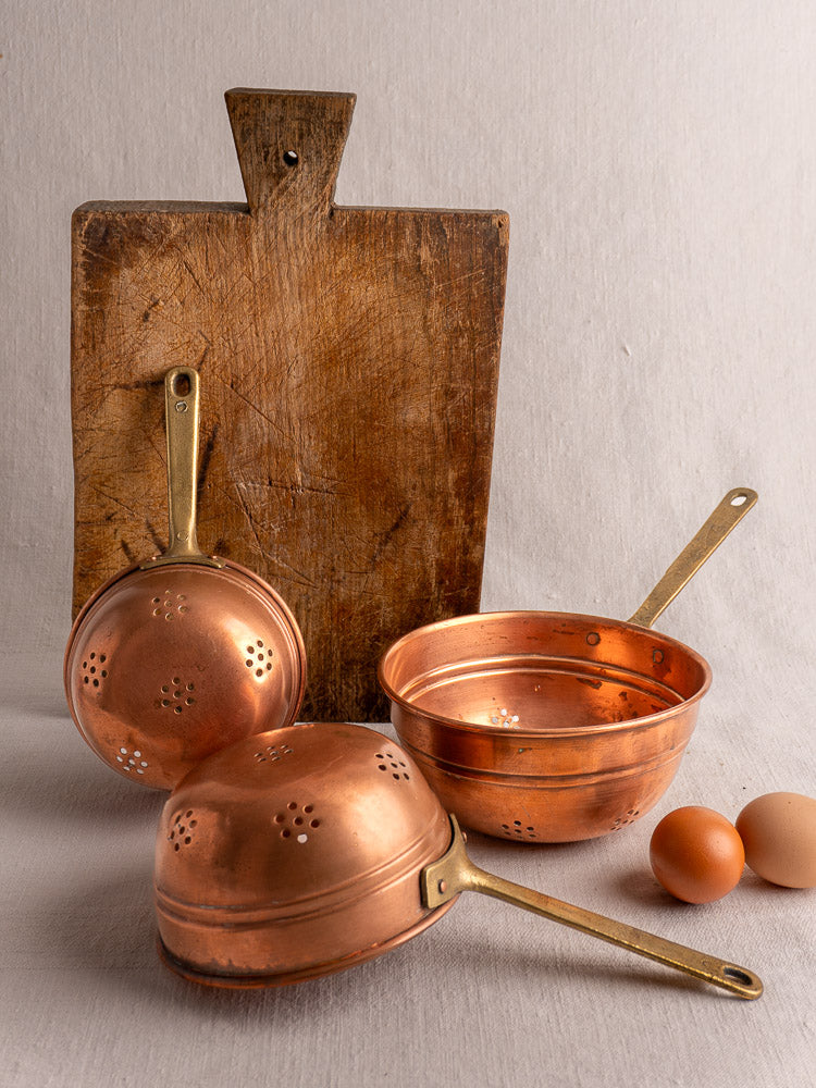 Antique French copper strainer set of three with brass handles, stacked on light beige fabric.
