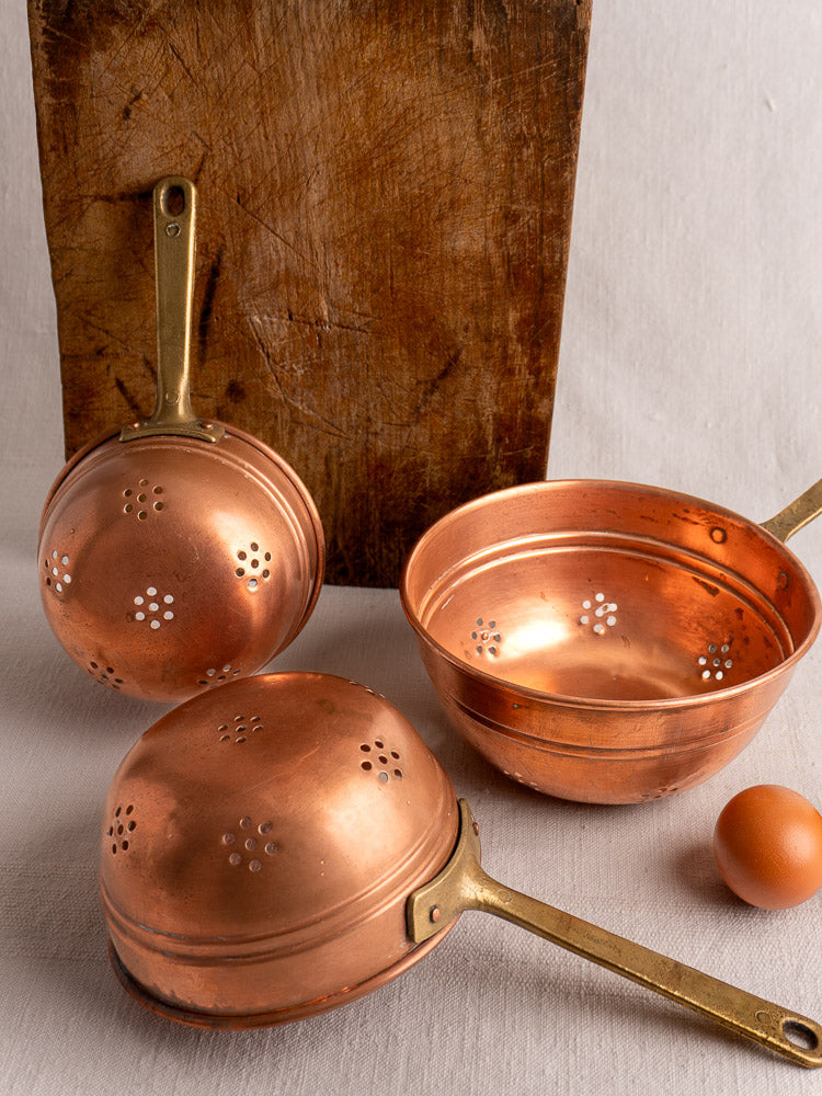 Antique French copper strainer set with brass handles, on a light surface with wooden board and brown egg nearby.