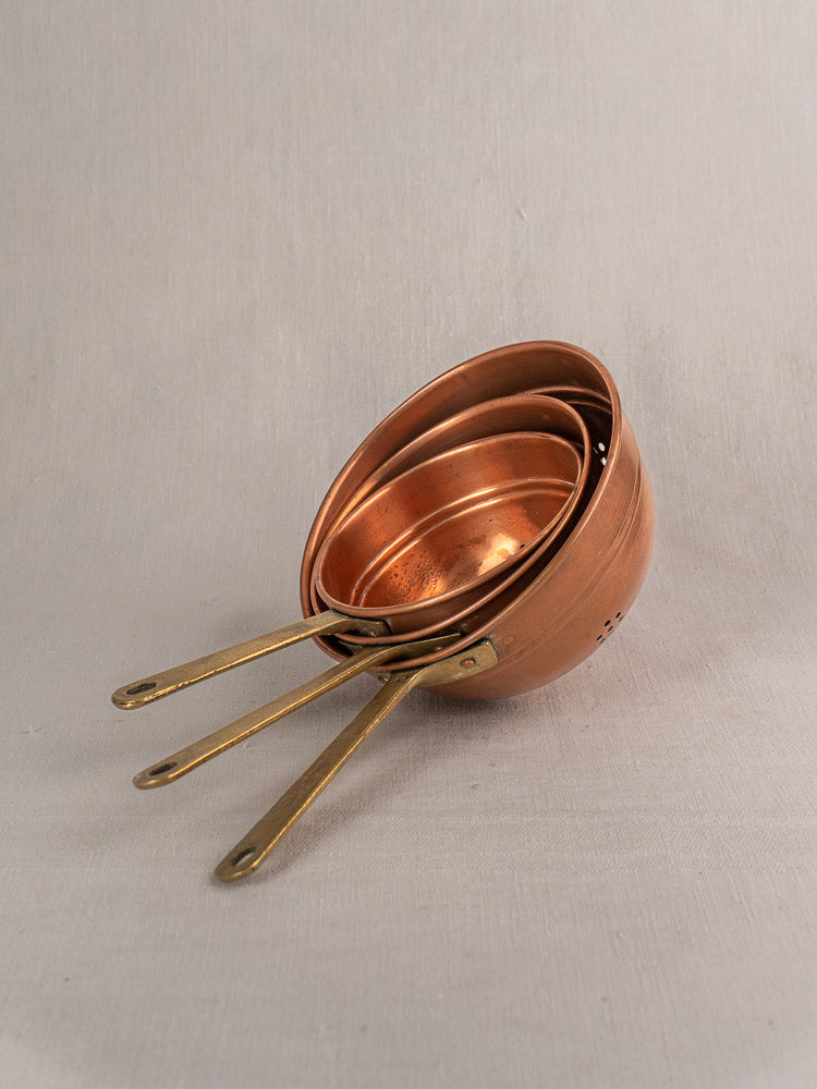 Antique French copper strainer set of three with brass handles, stacked on light beige fabric.