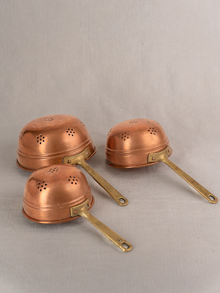 French antique copper strainer set of three colanders with brass handles, displayed upside down on neutral fabric.