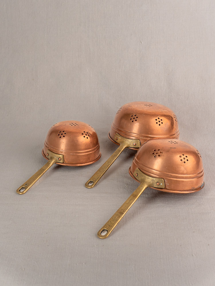 Set of three antique French copper colanders with brass handles, displayed upside down on a beige surface.