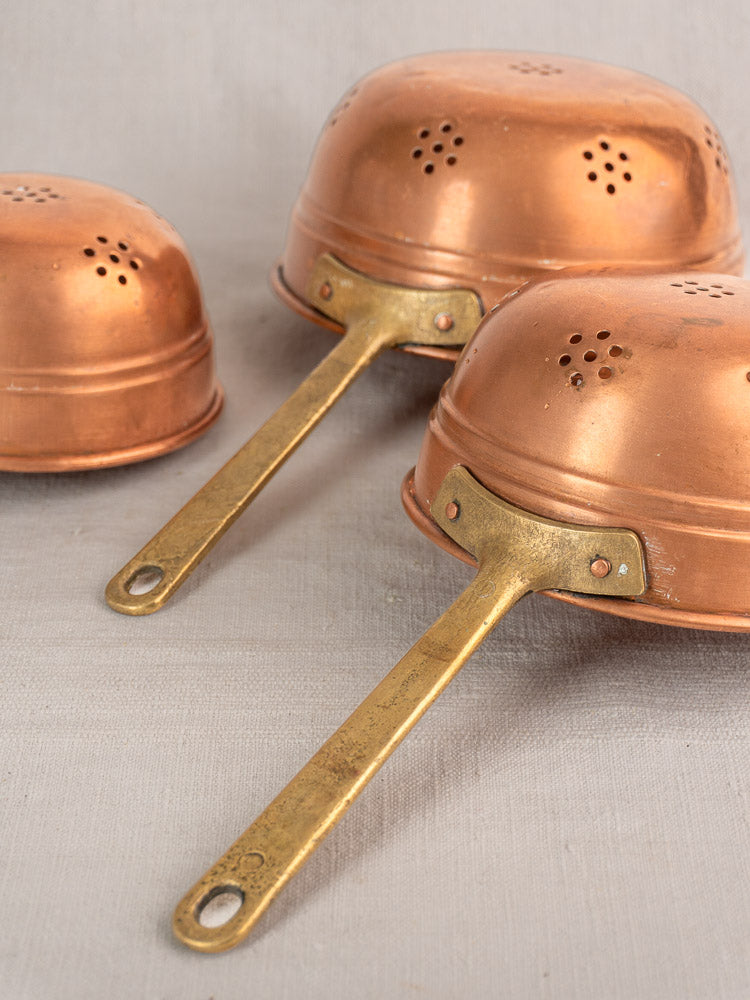 Antique French copper strainer set of three with brass handles, shown upside down on a light gray surface.