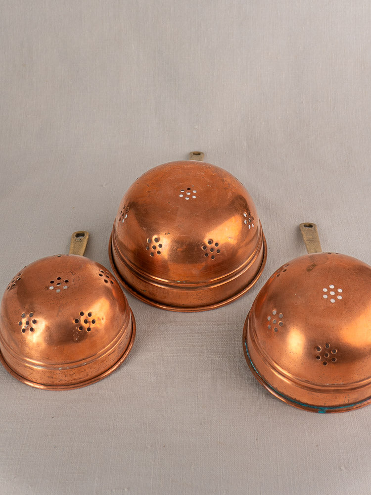 Antique French copper strainer set of three with brass handles, small perforations, on neutral fabric background.