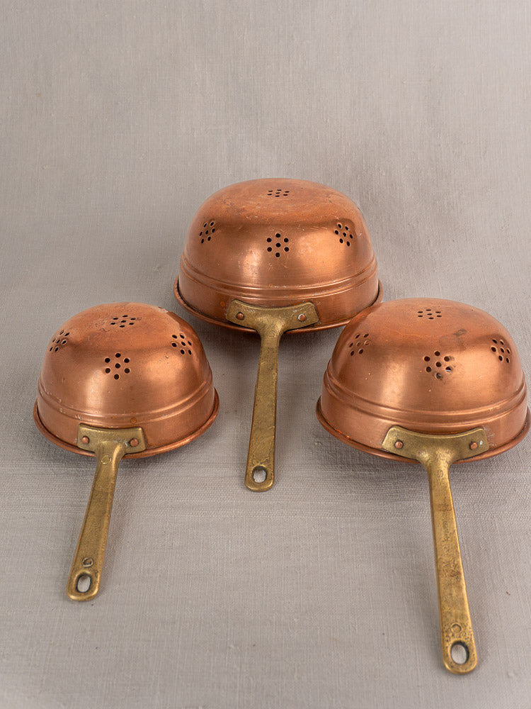 Antique French copper strainer set of three colanders with brass handles, decorative perforations, displayed upside down on gray.