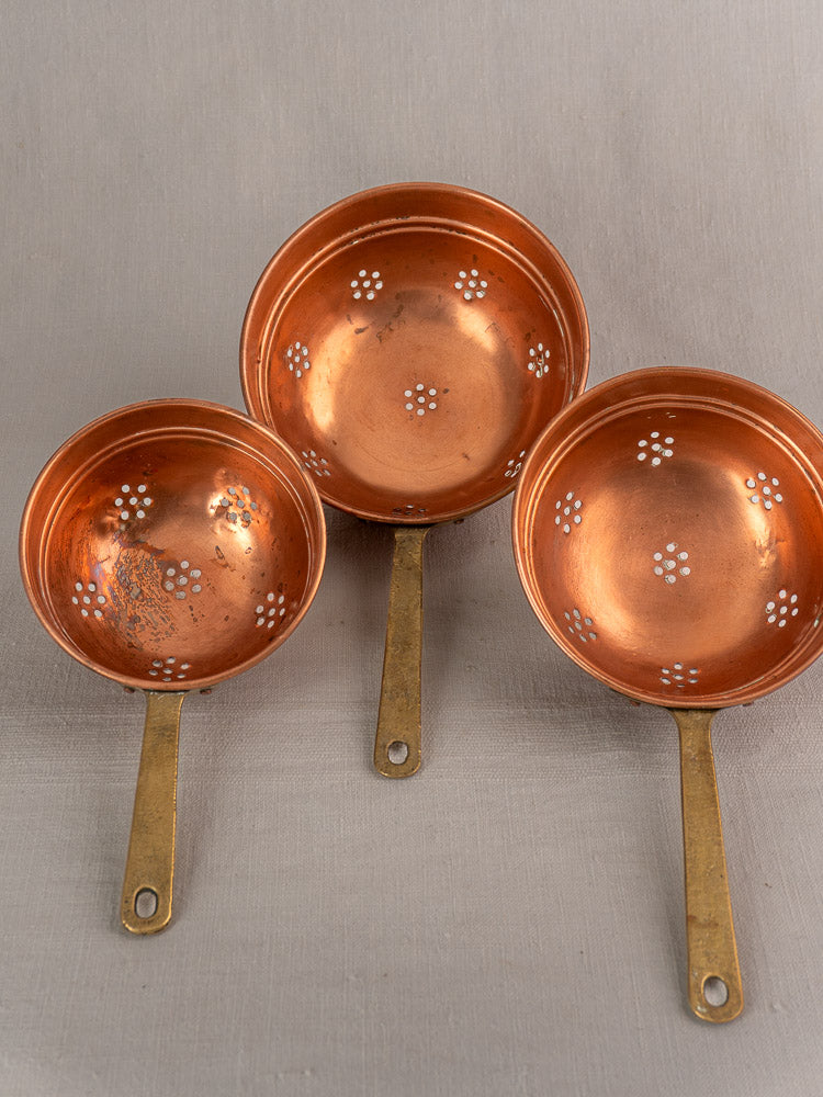 Antique French copper strainer set with brass handles, floral perforations, displayed on a light gray surface.
