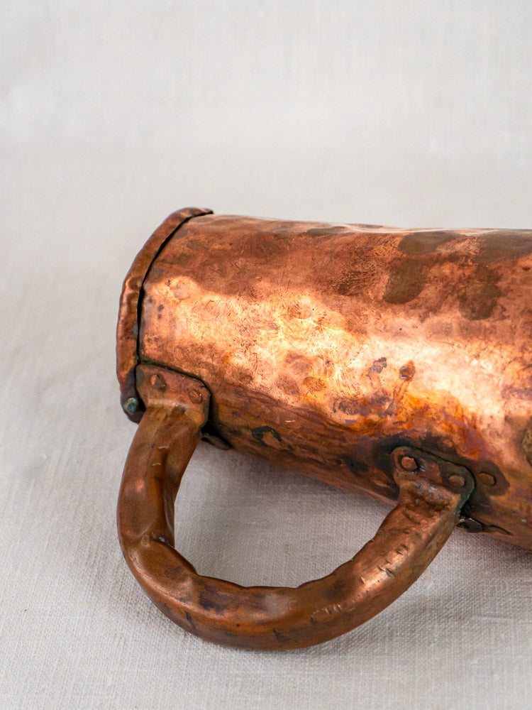 Antique French hand-forged copper chope tankard with hammered finish and curved handle on pale fabric.