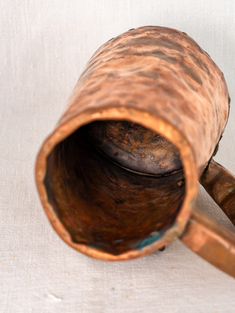 Antique French hand-forged hammered copper chope tankard with patina, dents, on light fabric background.