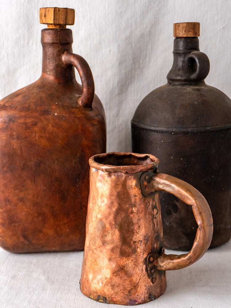Antique French copper tankard with hammered finish and large handle, flanked by rustic brown jugs with cork stoppers.