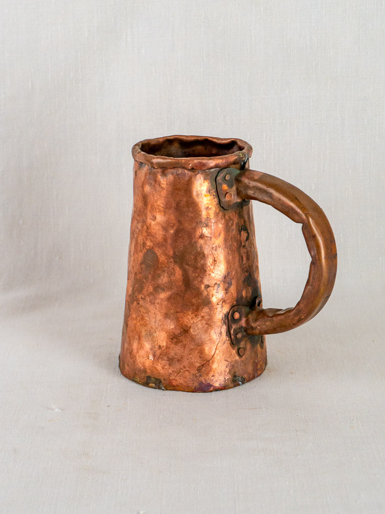 Antique French hand-forged copper tankard with large handle and hammer marks on light surface.