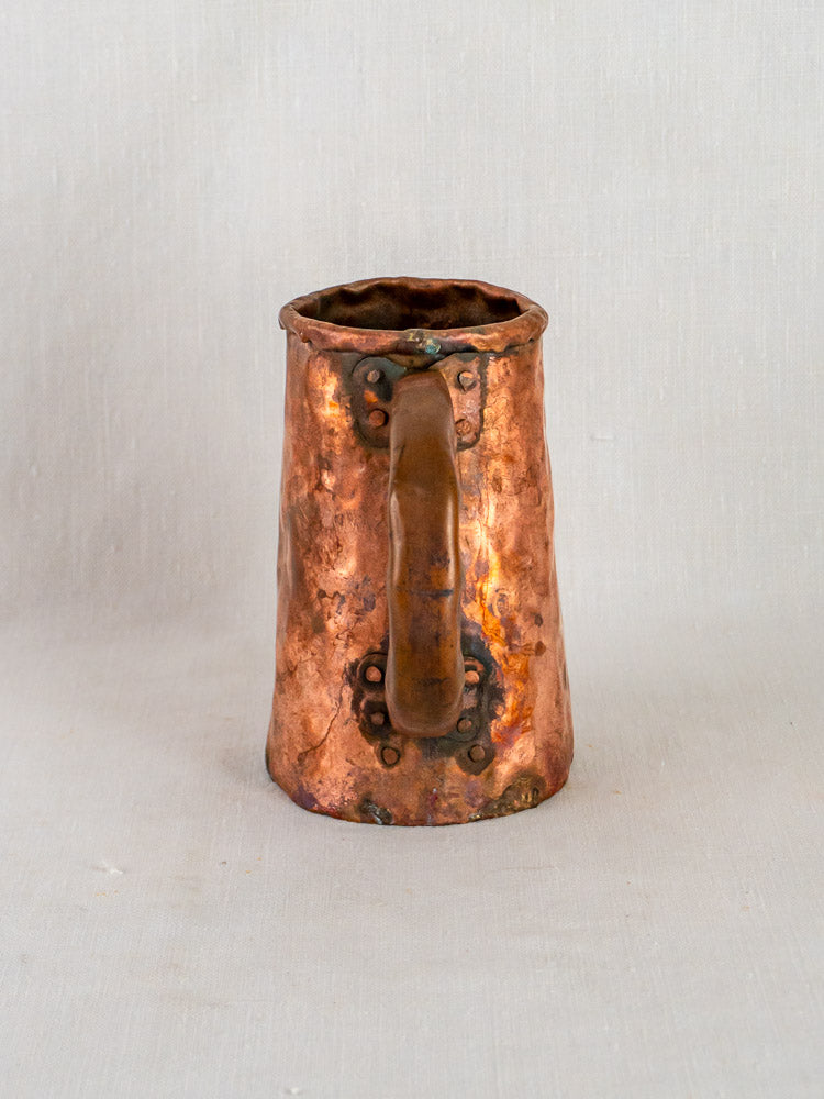 Antique French hand-forged copper tavern tankard with hammered finish, large handle, and riveted details on light fabric background.