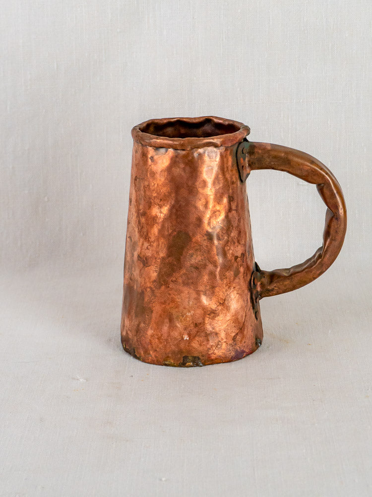 Hand-forged French copper tavern tankard with hammered texture and large handle on neutral surface.
