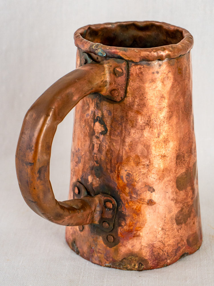Antique French copper chope tavern tankard with large handle, riveted joints, and aged patina on light backdrop.