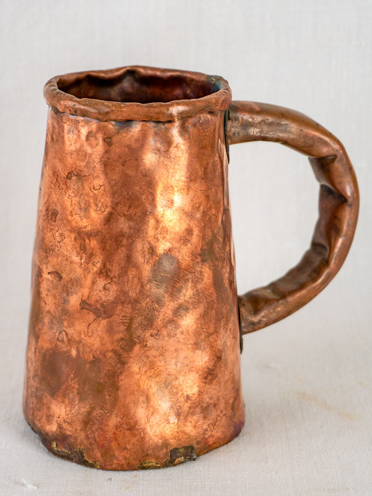 Antique French hand-forged copper tankard with hammered finish and curved handle on light background.