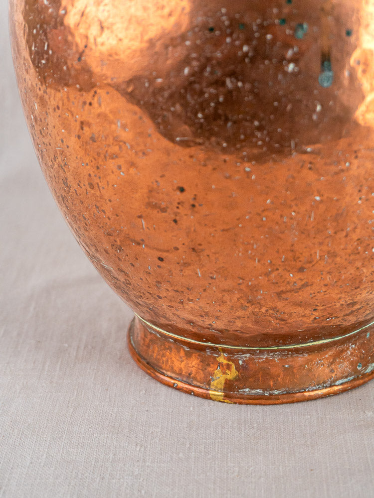 French hand-raised copper water jug with double spouts, tarnished base shows yellowish repair, on beige fabric.