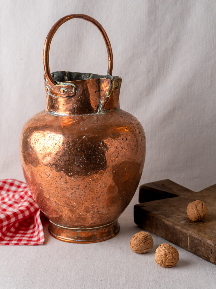 Antique French double-spout hammered copper water jug with handle, on cloth beside red gingham napkin and walnuts.