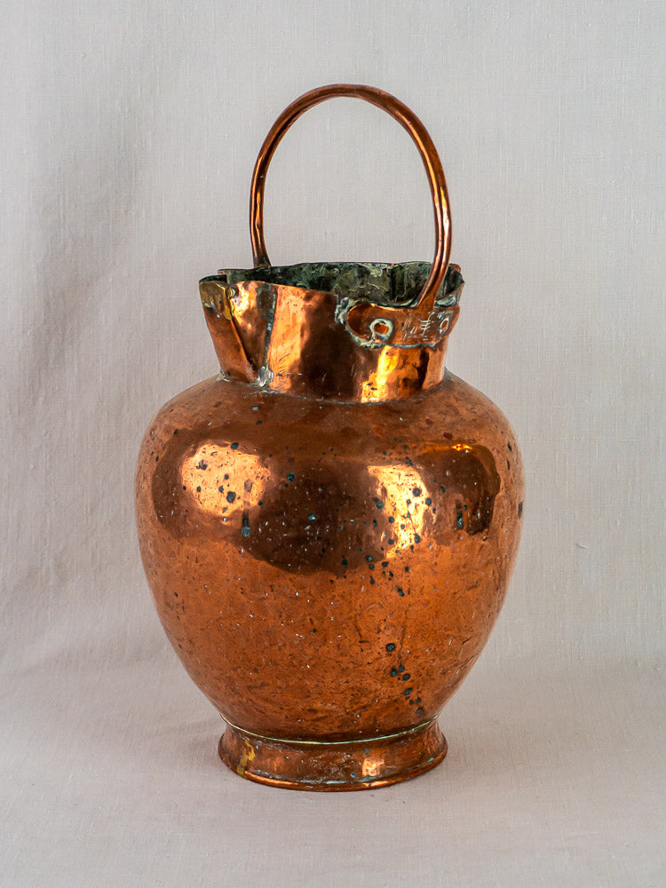 Antique French copper water jug with double spout, scalloped rim, and curved handle on fabric background.