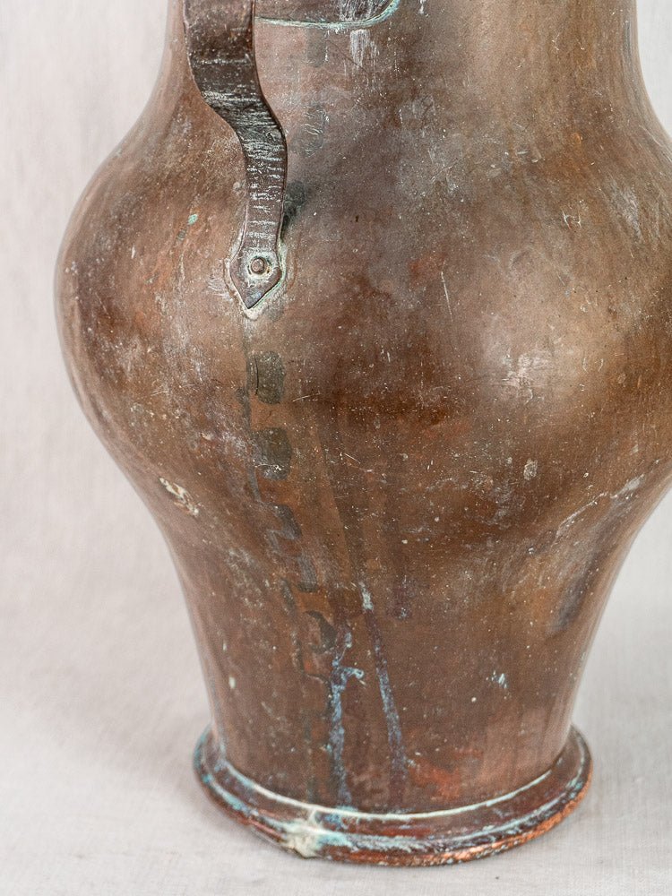 Antique French copper water jug with curved side, handle detail, dents, and green oxidation.