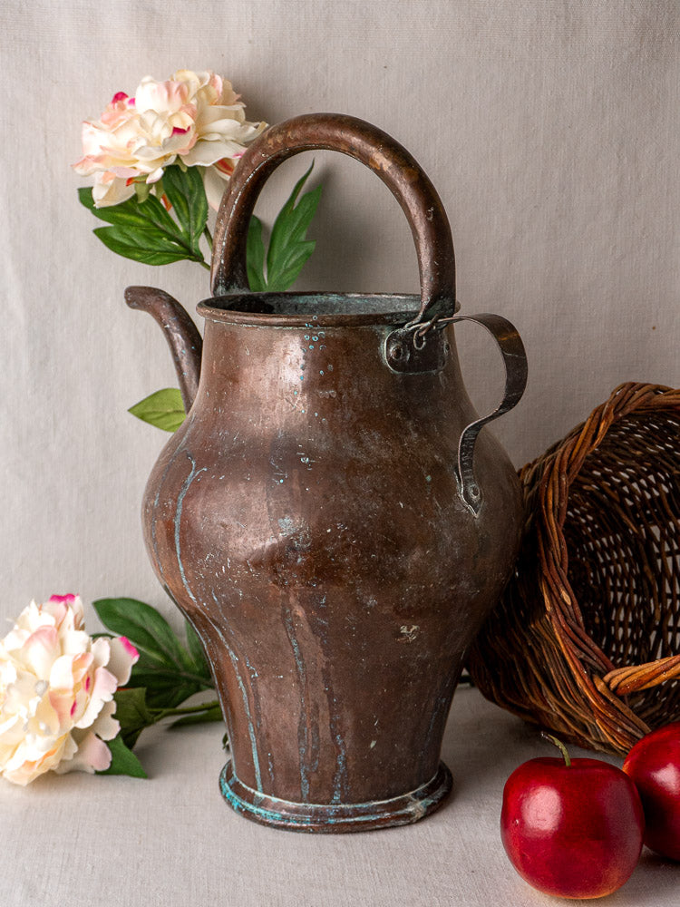 19th-century French copper water jug with wide base, curved spout, blue-green patina, on fabric backdrop.