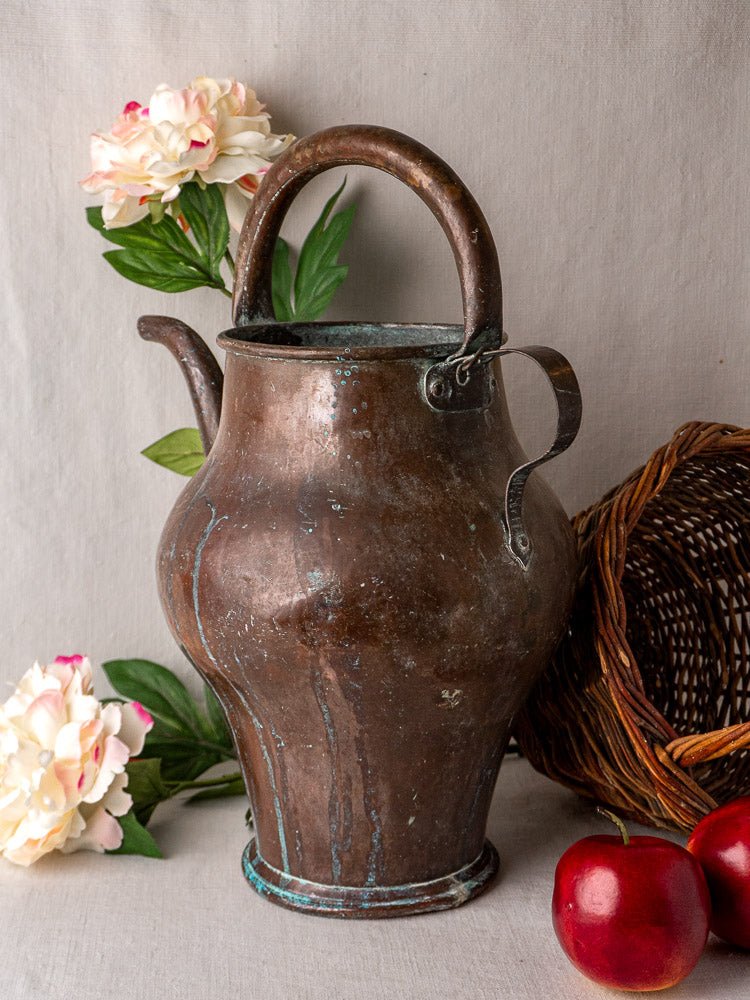Antique French copper water jug with curved handle, displayed beside basket, apples, and white flowers.