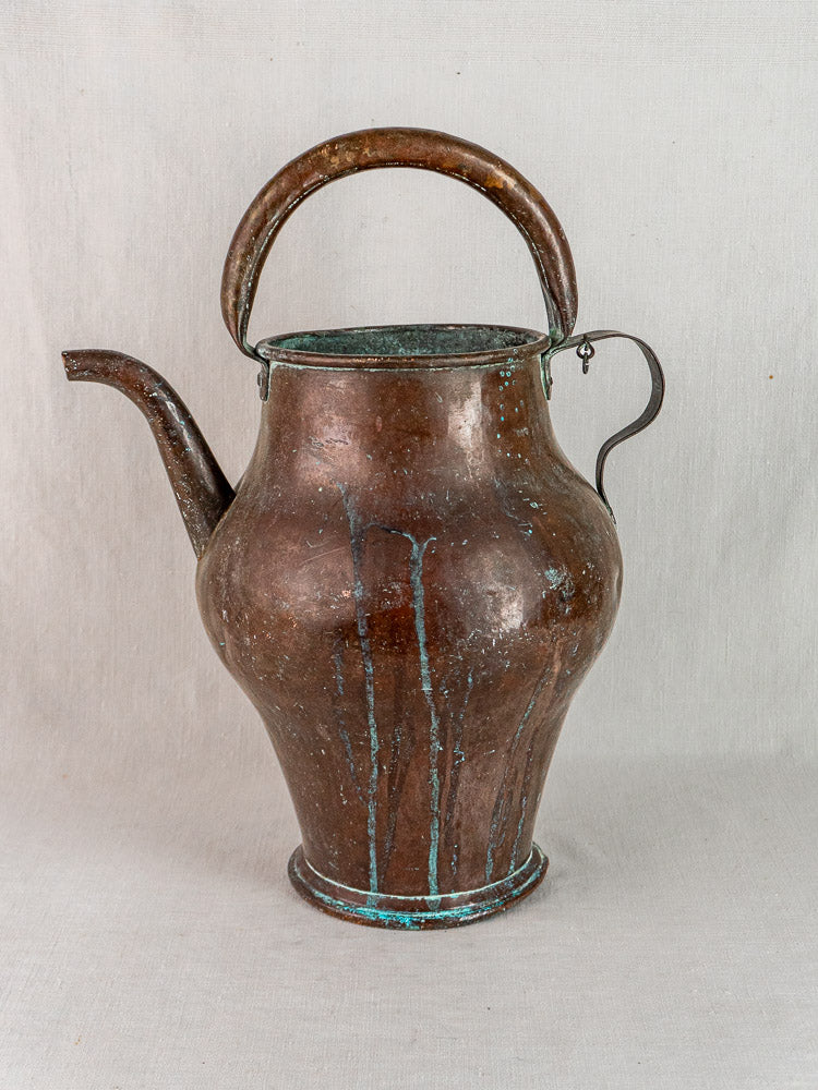 19th-century French copper water jug with wide base, curved spout, blue-green patina, on fabric backdrop.