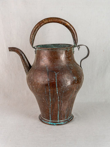 19th-century French copper water jug with wide base, curved spout, blue-green patina, on fabric backdrop.