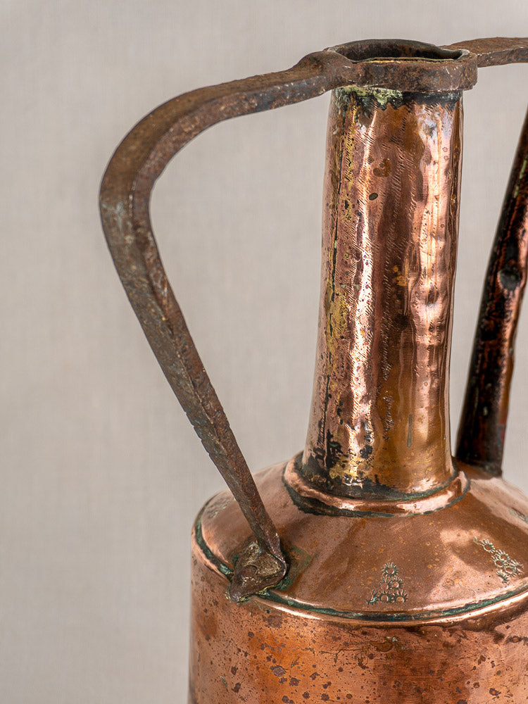 Antique French hand-hammered copper water jug with tall neck, curved iron handles, and rich patina on beige background.