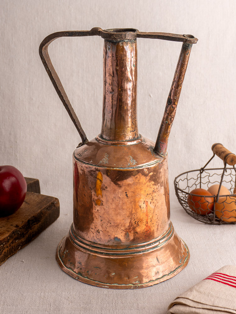 Antique French copper water jug with iron handles on linen, beside eggs, red apple, and wooden board.